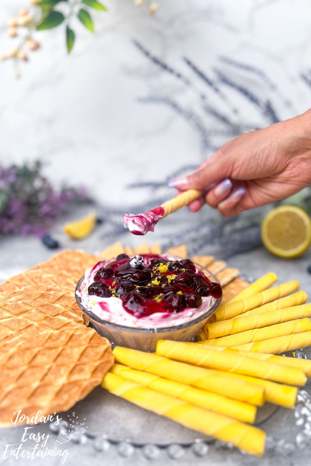 a lemon rolled wafer after scooping up a creamy cool blueberry cheesecake dessert dip topped with blueberry pie filling a lemon zest served with graham crackers and butter wafer cookies