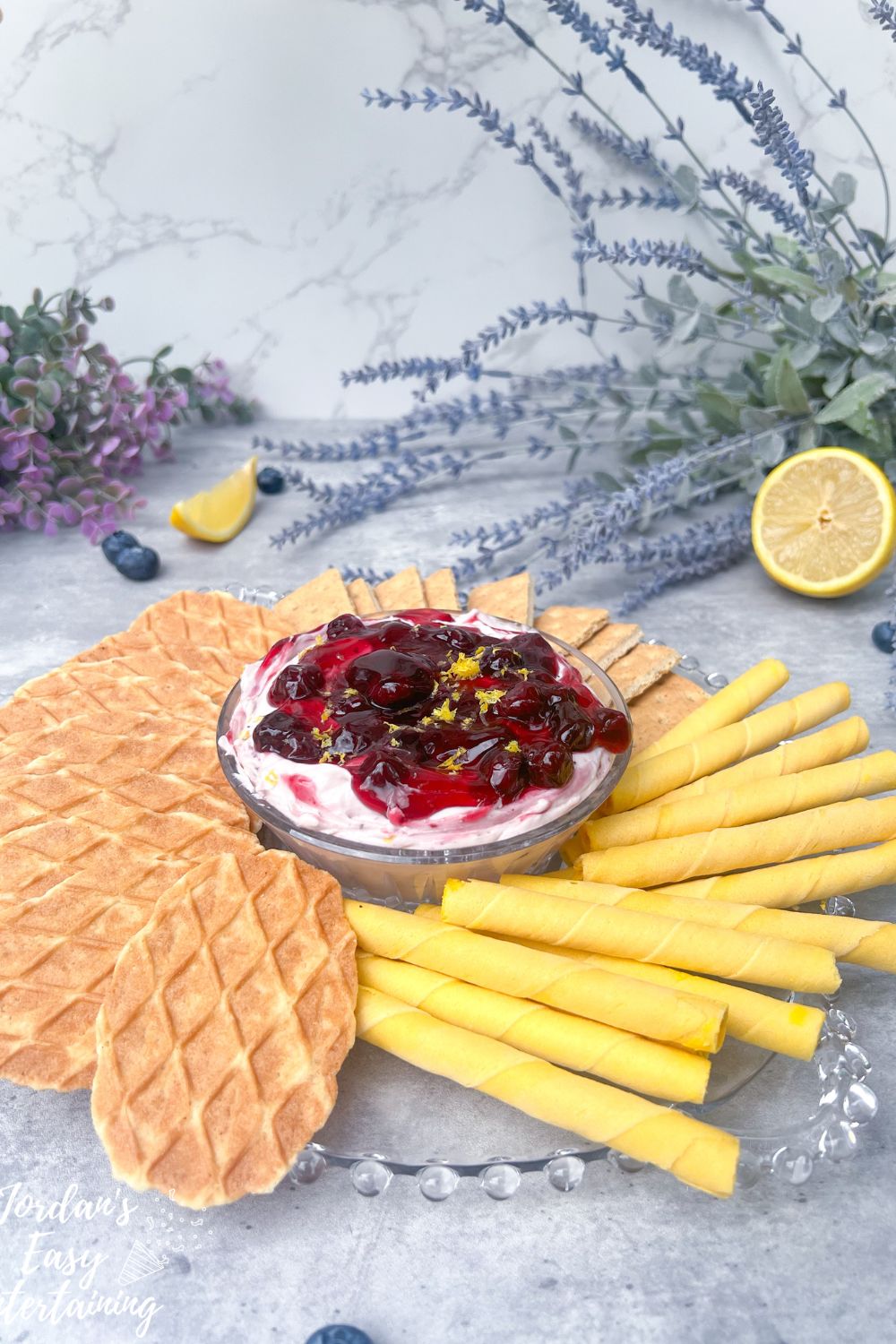 a bowl of cream cheese dessert dip topped with blueberry pie filling and lemon zest and served with lemon rolled wafers, butter wafer cookies, and graham crackers